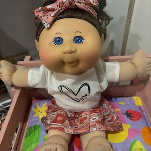 May include: A Cabbage Patch Kid doll wearing a pink and white headband, a white t-shirt with a black heart and the word "love" written on it, and a pink skirt with a heart pattern. The doll is sitting on a colourful bed with a pink headboard.