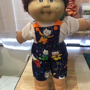 May include: A Cabbage Patch Kid doll wearing a white shirt and blue dungarees with a space cat print. The dungarees have orange straps and buttons.