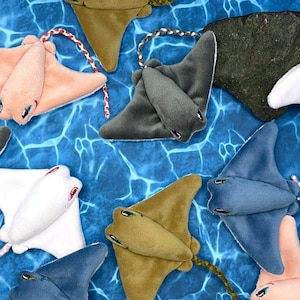 May include: A collection of plush stingray toys in various colors, including pink, white, gray, blue, and brown. The toys are arranged on a blue background with a textured pattern.