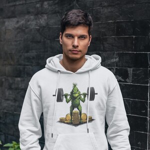 Può includere: Felpa con cappuccio bianca con un personaggio dei cartoni animati del Grinch che solleva un bilanciere. Il Grinch è verde e si trova su una pila di monete d'oro. La felpa ha una tasca frontale e un cappuccio con coulisse.