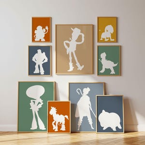 Könnte beinhalten: Eine Sammlung gerahmter Kunstdrucke mit weißen Silhouetten von Zeichentrickfiguren vor verschiedenen farbigen Hintergründen. Die Rahmen haben goldfarbene Ränder und sind an einer weißen Wand über einem Holzboden angeordnet.