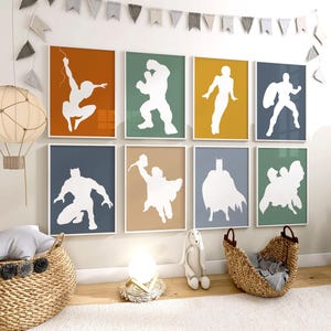 May include: A set of eight framed superhero silhouette art prints. Each print features a white superhero figure against a solid-color background, including orange, green, yellow, and blue. The prints are arranged on a wall above a wicker basket and other nursery decor.