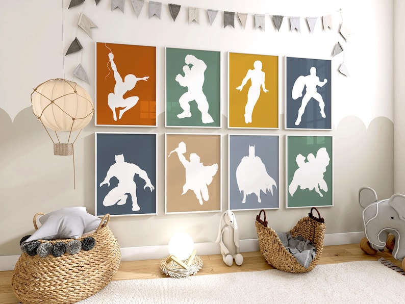 May include: A set of eight framed superhero silhouette art prints. Each print features a white superhero figure against a solid-color background, including orange, green, yellow, and blue. The prints are arranged on a wall above a wicker basket and other nursery decor.
