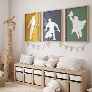 May include: Three framed superhero art prints in yellow, navy, and green. Each print features a white silhouette of a superhero. Below the prints is a wooden storage unit with white and beige storage bins. A woven giraffe and pillows complete the room.