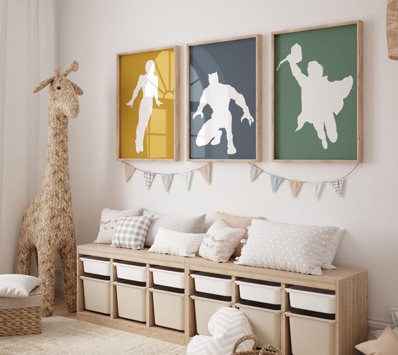 May include: Three framed superhero art prints in yellow, navy, and green. Each print features a white silhouette of a superhero. Below the prints is a wooden storage unit with white and beige storage bins. A woven giraffe and pillows complete the room.