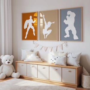 May include: Three framed superhero art prints with white silhouettes against orange, tan, and blue backgrounds. The prints hang above a wooden storage bench with white drawers and decorative pillows. A white teddy bear sits to the left.
