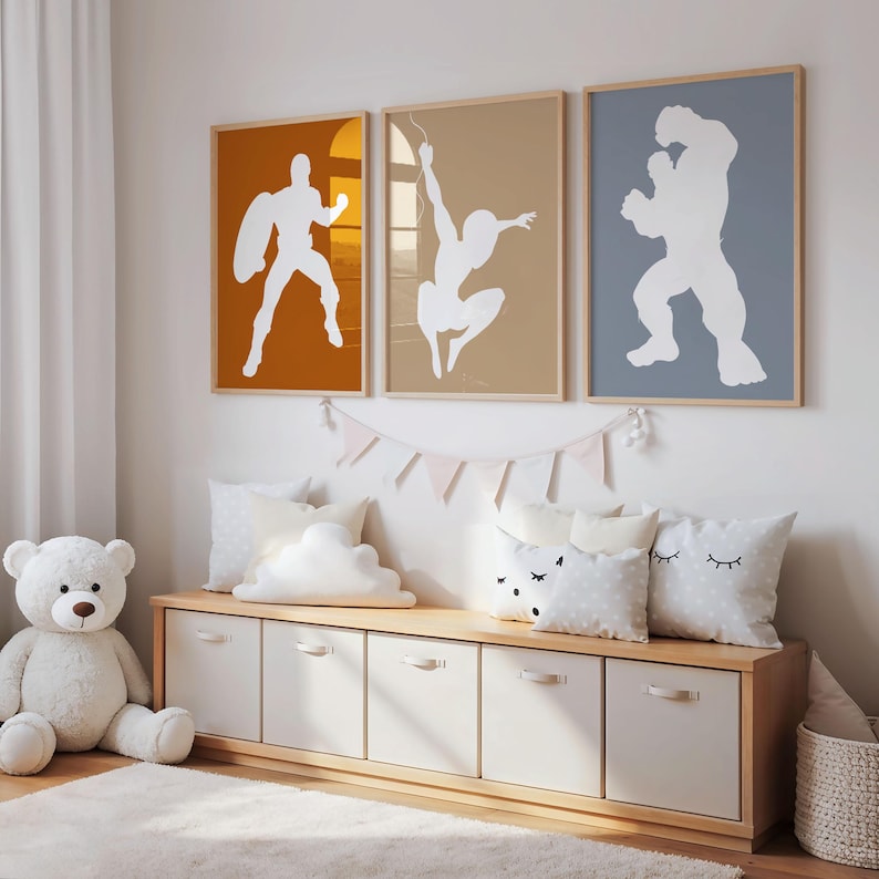 May include: Three framed superhero art prints with white silhouettes against orange, tan, and blue backgrounds. The prints hang above a wooden storage bench with white drawers and decorative pillows. A white teddy bear sits to the left.