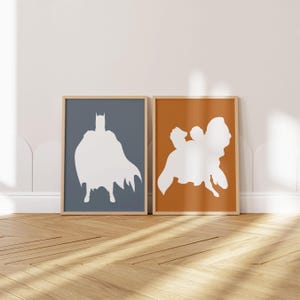 May include: Two framed art prints featuring white silhouettes against colored backgrounds. One print shows a Batman figure on a blue background, and the other depicts a figure on an orange background. The frames are a light brown color.