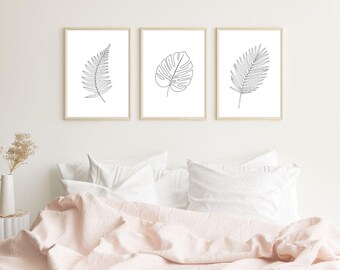May include: Three framed botanical art prints featuring line drawings of leaves. The prints are in light wood frames and hang above a bed with white bedding and a pink comforter. The leaves are a fern, a monstera, and a palm frond.