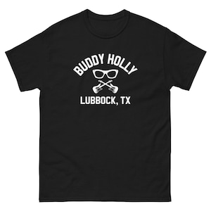 Può includere: Maglietta nera con testo bianco che recita "Buddy Holly Lubbock, TX" con un paio di occhiali e due chitarre incrociate.
