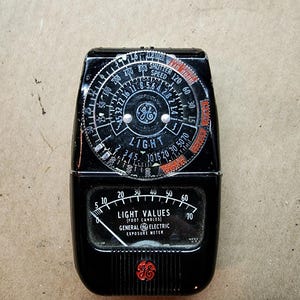 May include: A black General Electric exposure meter with a circular dial and a needle. The dial has markings for shutter speed, aperture, and light values. The meter has a red and white logo on the front.