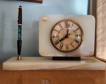 Könnte beinhalten: Ein weißer Marmor-Uhren- und Stifthalter mit einem goldfarbenen Zifferblatt und einem schwarzen Stift. Das Zifferblatt der Uhr hat römische Ziffern und die Worte "Imported Onyx" sind auf den Marmorsockel gedruckt.