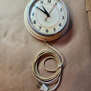 Könnte beinhalten: Eine weiße Vintage-Elektro-Uhr mit schwarzen Zeigern und einem schwarzen Rand. Das Zifferblatt der Uhr hat schwarze Zahlen und ein silbernes 'Westclox'-Logo. Die Uhr ist an eine Steckdose mit einem weißen Kabel angeschlossen.