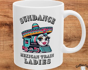 May include: White ceramic mug with a colorful cartoon train wearing a sombrero. The text "SUNDANCE MEXICAN TRAIN LADIES" is printed on the mug.
