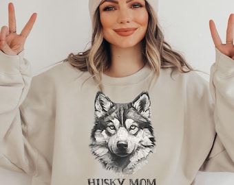 May include: Beige sweatshirt featuring a detailed black and white illustration of a husky dog's face. The text "HUSKY MOM" is printed below the image in a bold, black font. The sweatshirt is a neutral color, suitable for casual wear.