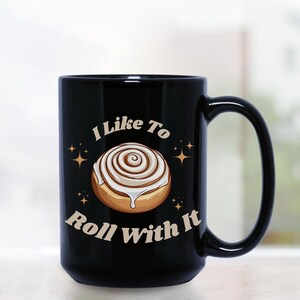 May include: Black coffee mug with a white cinnamon roll graphic and the text "I Like To Roll With It".