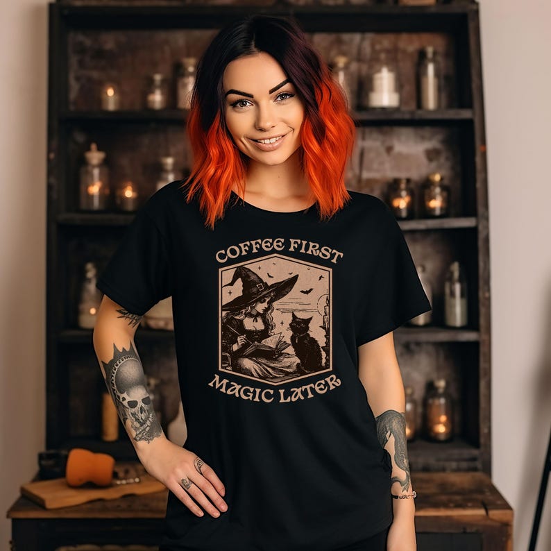 May include: Black t-shirt with a vintage-style illustration of a witch reading a book with a black cat. The text "Coffee First Magic Later" is printed on the shirt.