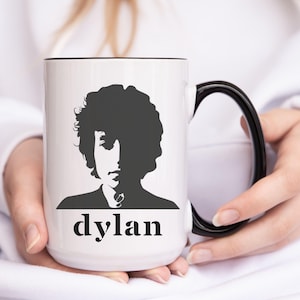 May include: White ceramic mug with a black handle. The mug features a black and white silhouette of Bob Dylan with the word "dylan" printed below the image.