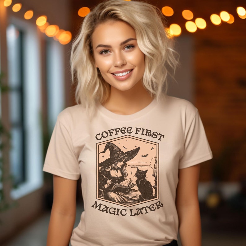 May include: A beige t-shirt with a vintage-style illustration of a witch reading a book with a black cat beside her. The text "Coffee First Magic Later" is printed on the shirt.