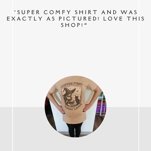 May include: A tan t-shirt with a black and white illustration of a witch and a cat. The text "Coffee First Magic Later" is printed on the shirt.