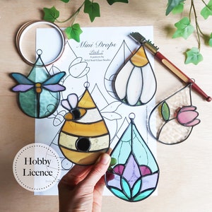 May include: Five stained glass mini drop ornaments in various designs, including a dragonfly, a beehive, a flower, and a white flower. The ornaments are on a white sheet of paper with a black outline of each ornament. The paper has the text "Mini Drops Pack 2 A pattern by Wild Soul Glass Studio" printed on it.