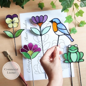 Può includere: Una collezione di colorati segnaposto per piante in vetro colorato, tra cui una viola, un fiore viola, un uccello blu e una rana verde. I segnaposto sono montati su sottili aste di metallo. Sullo sfondo un foglio con la scritta "Cottage Plant Stakes".