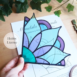 Könnte beinhalten: Ein Buntglas-Mandala in Blau-, Lila- und Grüntönen mit schwarzem Rand. Das Design ist floral und geometrisch, mit dem Text "Mandala" und "A pattern by Wild" auf dem Papier darunter. Das Hobby Licence-Logo ist ebenfalls sichtbar.