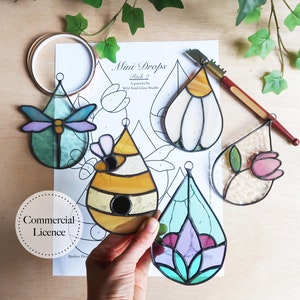 May include: Five stained glass window hangings in the shape of teardrops, each with a different design. The designs include a dragonfly, a beehive, a flower, a flower with a bee, and a flower with a butterfly. The hangings are on a white paper with a black outline of each design.