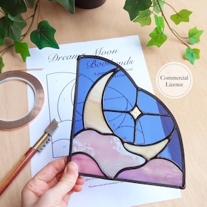 Könnte beinhalten: Ein Buntglasstück mit einem Mond, Stern und Wolken in Blau-, Rosa- und Cremetönen. Das Kunstwerk wird über einem Muster mit den Worten "Dream Moon Bookends" gehalten. Eine Kupferfolienrolle und ein Glasschneider sind ebenfalls zu sehen.