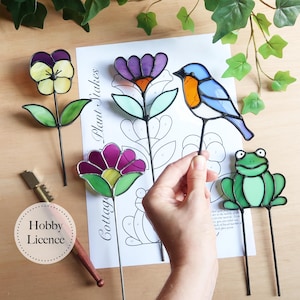 Könnte beinhalten: Eine Sammlung farbenfroher Glasmalerei-Pflanzstäbe, darunter ein Stiefmütterchen, eine lila Blume, ein blauer Vogel und ein grüner Frosch. Die Stäbe liegen auf einer Holzoberfläche mit einem Designblatt "Cottage Plant Stakes".