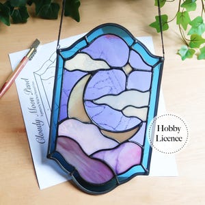 Puede incluir: Un panel de vidrieras con una luna creciente y nubes en tonos de morado, lavanda y crema. El panel está enmarcado en negro y tiene una cadena para colgar. El texto "Cloudy Moon Panel" es visible en un papel cercano.