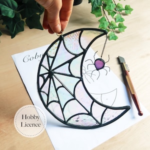 Puede incluir: Una luna creciente de vidriera con un diseño de telaraña y una araña morada colgando de una cadena. La luna es iridiscente con bordes negros. El texto "Hobby Licence" está en una pegatina.