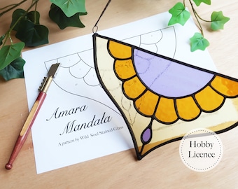 May include: A stained glass mandala in yellow, purple, and clear glass, with a black outline. The design is a half-circle with a petal-like border. A pattern and glass cutter are next to the mandala. The text "Amara Mandala" is visible.