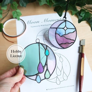Puede incluir: Dos piezas de vitral, una con un diseño de luna y estrellas en tonos de rosa, azul y blanco, y la otra con un diseño de lágrima en tonos de verde, azul y morado. Las piezas están sobre una hoja de papel blanco con un patrón impreso en ella. El texto "Moon Moments" y "A pattern by Wild Soul Stain" está impreso en el papel. El texto "Hobby Licence" está impreso en un círculo blanco.