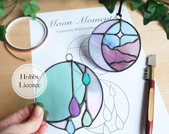 May include: Two stained glass pieces, one with a moon and stars design in shades of pink, blue, and white, and the other with a teardrop design in shades of green, blue, and purple. The pieces are on a white sheet of paper with a pattern printed on it. The text "Moon Moments" and "A pattern by Wild Soul Stain" is printed on the paper. The text "Hobby Licence" is printed on a white circle.