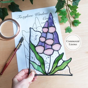May include: A stained glass window panel depicting a purple and pink foxglove flower with green leaves. The panel is being held by a hand and is on a wooden surface. The image also includes a pattern sheet and a copper tape roll.
