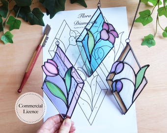 May include: Three diamond-shaped stained glass window hangings with floral designs. The hangings are in shades of blue, purple, and pink. The hangings are on a white sheet of paper with a pattern printed on it. The paper has the text "Flora Diamonds" printed on it.