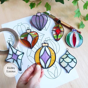 Può includere: Una collezione di ornamenti natalizi in vetro colorato di varie forme, tra cui cuori e stelle. Ogni ornamento presenta un design unico con colori vivaci e contorni neri. Gli ornamenti sono esposti su una superficie bianca con un motivo stampato.