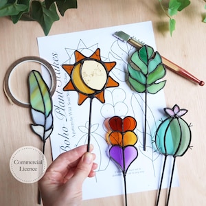 May include: A selection of stained glass plant stakes in the shapes of a sun, leaf, feather, heart, and cactus. The stakes are in orange, yellow, green, purple, and white. The text "Boho Plant Stakes" is visible on a paper pattern.
