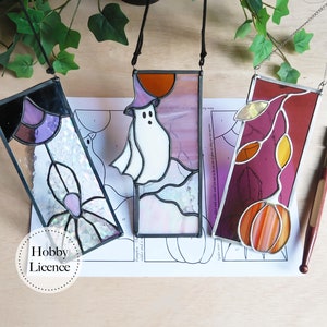 Op de afbeelding: Drie gebrandschilderde raamhangers met Halloween-thema's. De eerste toont een paarse en bruine spin, de tweede een witte geest met een pompoen en de derde een rode en oranje pompoen met bladeren.