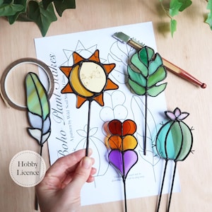 Puede incluir: Una colección de coloridas estacas para plantas de vidrieras en varias formas, incluyendo un sol, una hoja, una pluma, un corazón y un cactus. Las estacas están sobre varillas de metal negro. La imagen también muestra una hoja de patrones con el texto "Boho Plant Stakes".