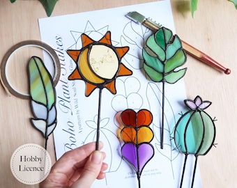 May include: A collection of colorful stained glass plant stakes in various shapes, including a sun, leaf, feather, heart, and cactus. The stakes are on black metal rods. The image also shows a pattern sheet with the text "Boho Plant Stakes".