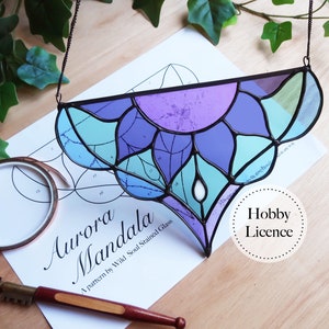 May include: A stained glass mandala with a purple, blue, and green color scheme. The mandala is hanging from a chain and is labeled "Aurora Mandala" and "A pattern by Wild Soul Stained Glass".