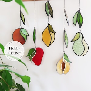 May include: A set of stained glass fruit ornaments hanging from chains. The ornaments include a peach, a lemon, a mango, a pear, and a quince. The ornaments are all different colors and have green leaves.