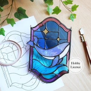 Könnte beinhalten: Buntglas-Kunstwerk mit einem Wal-Design in Blau- und Lilatönen. Das Kunstwerk hat einen dekorativen Rahmen in Fensterform. Im Hintergrund sind ein Muster und ein Glasschneider zu sehen.