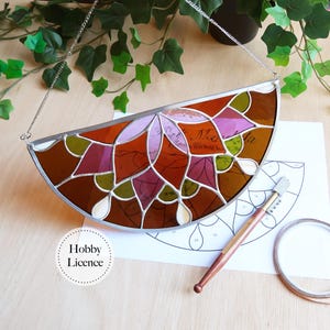 May include: A half-circle stained glass panel with a silver chain for hanging. The panel features a floral design in shades of brown, orange, purple, and green. A pattern and tools for creating stained glass are visible below the panel.