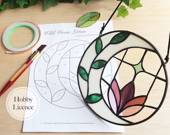 May include: A Wild Moon Lotus stained glass pattern with a Hobby Licence. The pattern is accompanied by a stained glass piece, copper tape, and a glass cutter. The stained glass piece features a floral design with green, white, and pink hues.