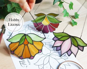 May include: Three stained glass flower suncatchers hanging from chains. The flowers are in shades of purple, pink, and orange with green leaves. The suncatchers are hanging over a sheet of paper with a flower pattern and the words "Mini Flower" printed on it.