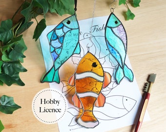 May include: Three stained glass fish hang from strings. The fish are blue, orange, and white. The fish are hanging over a sheet of paper with a fish outline and the word "Fish" printed on it. The paper also has the words "Hobby Licence" printed on it.