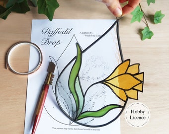 May include: A stained glass daffodil pattern, with a yellow flower and green leaves, on a white paper background. The pattern is titled "Daffodil Drop" and includes the text "A pattern by Wild Soul Glass" and "This pattern may not be distributed or sold in any way". The image also includes a hobby licence.